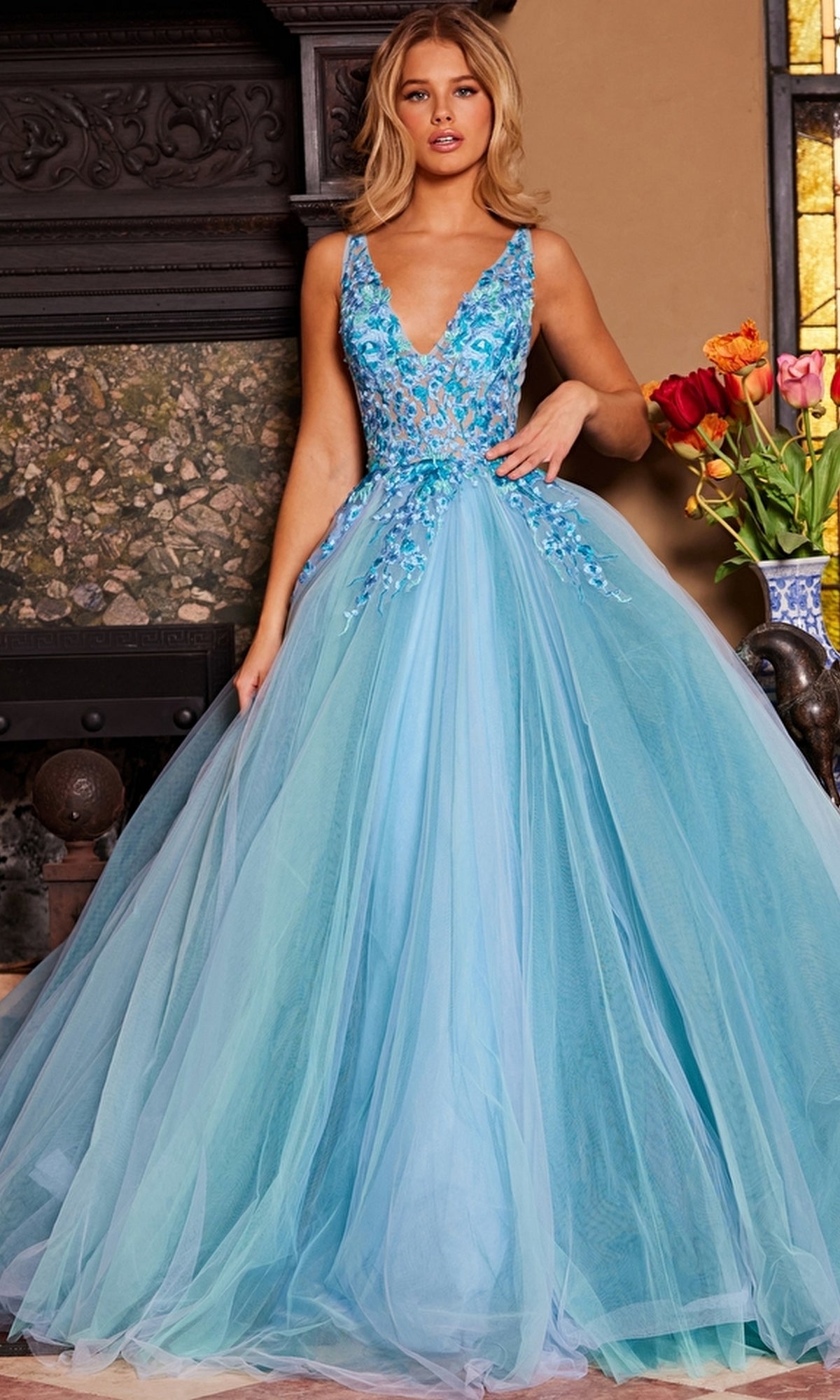 A woman stands indoors in the Jovani Tulle Prom Ball Gown with Floral Embroidery, a floor-length, sleeveless dress featuring a flowing tulle skirt. A vase of flowers and a fireplace accent the elegant setting.